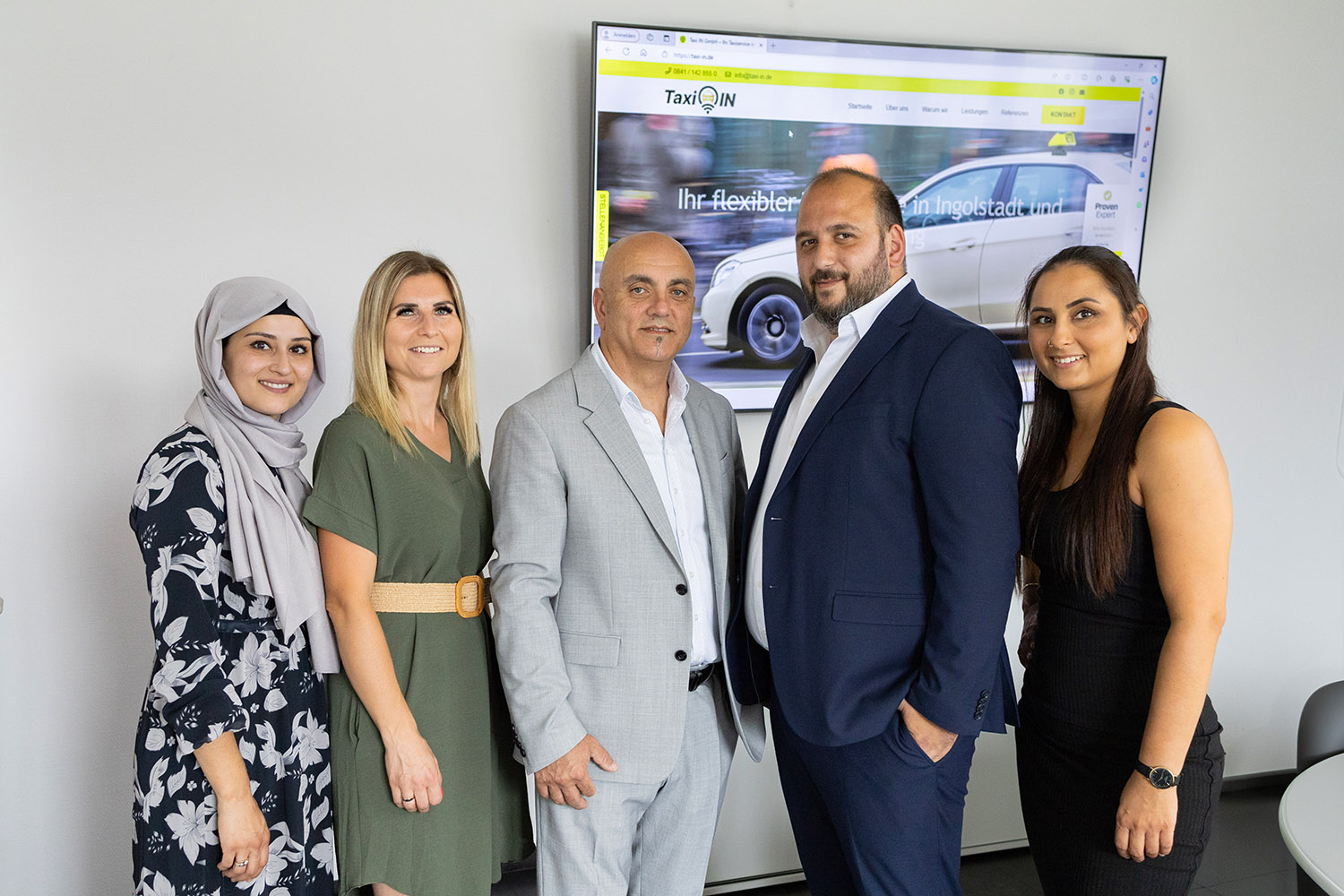 Fünf Personen stehen nebeneinander in einem Büroraum vor einem großen Bildschirm mit der Taxi-IN-Website im Hintergrund.
