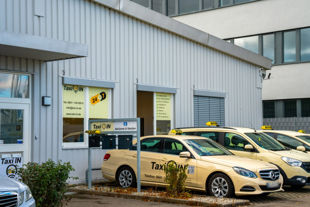 Cremefarbene Taxi-Flotte vor dem Taxi-IN-Firmengebäude mit Beschilderung, Bürofenstern und mehreren geparkten Fahrzeugen.