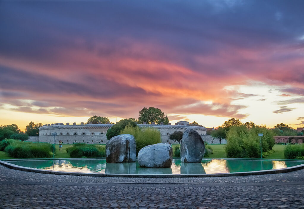 Springbrunnen mit großen Felsblöcken im Klenzepark Ingolstadt bei dramatischem Sonnenuntergang, im Hintergrund die historische Reduit-Tilly-Anlage.